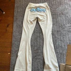 Vita Juicy Couture mjukisbyxor XXS - Vita mjukisbyxor från Juicy Couture i supermjuk velour med blått broderat Juicy-tryck och blomma bak. Byxorna har snörning i midjan och JC-logga fram. De har bootcut-modell och är perfekta för en chill och trendig look.