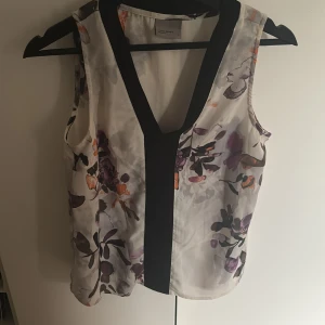 Blommig ärmlös blus från Vero Moda - Snygg ärmlös blus från Vero Moda i vitt med blommigt mönster i lila, svart och orange. Toppen har en djup v-ringning med svart kant framtill och är tillverkad i ett lätt, flowy material som känns luftigt och fräscht.