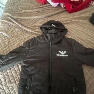 Svart Emporio Armani pufferjacka - Svart pufferjacka från Emporio Armani med huva och dragkedja framtill. Jackan har logga tryckt på bröstet och på huvan, samt två fickor med dragkedja. Perfekt för kalla dagar när du vill hålla stilen. Inget e sönder den e i bra kvalitet allt e bra med jackan