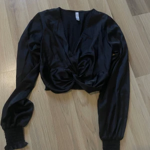 Svart omlottopp i satin-look - Snygg svart omlottopp i glansigt satinliknande material med croppad passform. Toppen har långa puffiga ärmar med breda smockade muddar och en twistad detalj framtill. Perfekt för dig som gillar trendiga och stilrena plagg med det lilla extra.
