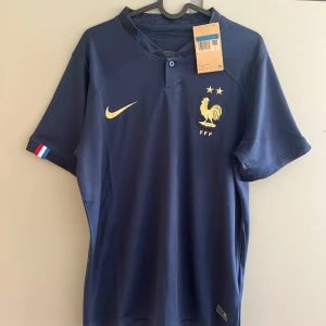 Frankrike fotbollströja Nike blå - Snygg marinblå Frankrike fotbollströja från Nike med guldiga detaljer, FFF-logga och två stjärnor på bröstet. Kortärmad modell med liten krage och knapp framtill. Fransk flaggdetalj på ärmslutet. Perfekt för dig som älskar landslagsfotboll.