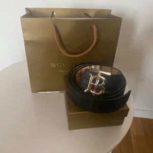 Burberry bälte med guldspänne - Säljer ett Burberry bälte med ikoniskt rutigt mönster i beige, rött och brunt. Bältet har ett stort guldfärgat 'TB'-spänne och är tillverkat i skinn med textil på utsidan. Perfekt accessoar för att lyfta din outfit med klassisk Burberry-stil.