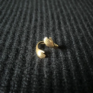 Câlin Doré – Golden Hug Ring - Justerbar ring i rostfritt stål med guldplätering, rost- och nickelfri. Två armar och händer sveper runt fingret i en omfamnande form – en stilren design med subtil närhet