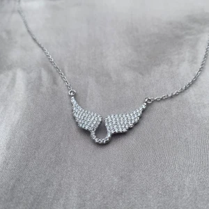 Silverhalsband med glittriga vingar - Ett snyggt halsband i silverfärg med ett hänge format som två vingar täckta av glittrande stenar. Kedjan är tunn och smidig med ett klassiskt lås. Perfekt för dig som vill ha något unikt och iögonfallande med lite extra bling.