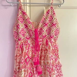 Mönstrad rosa och vit klänning - Superfin kortklänning med tunna axelband, v-ringning och broderade detaljer i rosa, vitt och guld. Klänningen har ett färgstarkt mönster, volanger och snörning med tofsar framtill. Perfekt för sommarens varma dagar.