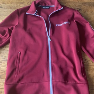 Röd track jacket från Palm Angels - Snygg röd track jacket från Palm Angels med vita ränder längs ärmarna och logga på bröstet och ryggen. Jackan har dragkedja framtill, ståkrage och två fickor. Perfekt för en streetwear-look.