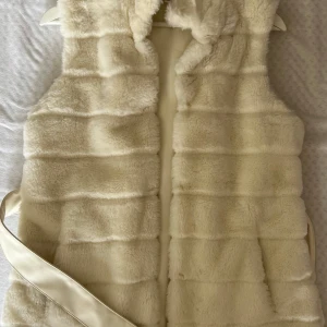 Krämvit pälsväst från New Yorker med bälte - Supermjuk och fluffig krämvit pälsväst från Amisu i storlek 36. Västen har horisontella sömmar för en cool struktur och ett matchande bälte i satinliknande material som knyts i midjan. Perfekt för lager-på-lager och att lyfta din outfit.