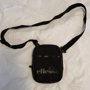 Svart axelväska från Ellesse - Säljer en svart axelväska från Ellesse med justerbar rem och dragkedja framtill. Väskan har en kompakt, fyrkantig form och är perfekt för att bära det viktigaste. Stort Ellesse-tryck på framsidan ger en sportig vibe.