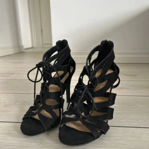 Svarta klackar med snörning - Snygga svarta sandaler med hög klack och öppen tå. Skorna har trendiga utskurna detaljer, snörning framtill och dragkedja bak för enkel påtagning. Tillverkade i mocka-look som ger en lyxig känsla och passar perfekt till en dressad outfit.