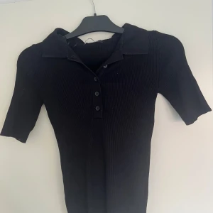Svart ribbad polotopp från Gina Tricot - Svart ribbad polotopp från Gina Tricot med korta ärmar och klassisk krage. Toppen har knappar framtill och är figurnära i modellen. Perfekt för dig som gillar en stilren och enkel look med lite edge. Kontakta för fler bilder eller frågor!