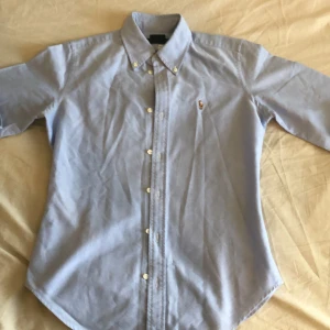 Ljusblå skjorta från Ralph Lauren - Klassisk ljusblå skjorta från Ralph Lauren i super slim fit. Skjortan är i 100% bomull, har långa ärmar, knappar framtill och button down-krage. Ikoniska broderade loggan på bröstet. Perfekt för en clean och stilren look.