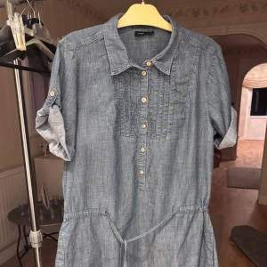 Blå kort skjortklänning från Lindex - Snygg blå skjortklänning i  från Lindex. Modellen är kort med knappar framtill, krage och knytband i midjan. Ärmarna är trekvarts och kan rullas upp och fästas med knapp. Praktiska fickor på sidorna.  Det står storlek 44 men den passar utan tvekan någon mindre också.