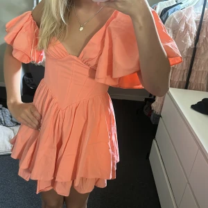 Persikofärgad kortklänning med volang - Supersöt kortklänning i persikofärg med offshoulder och stora volangärmar. Klänningen har en markerad midja, omlottkänsla upptill och en fluffig, dubbellagrad kjol. Perfekt för dig som vill sticka ut med en färgglad och lekfull look.