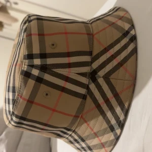 Burberry rutig bucket hat beige M - Säljer en klassisk bucket hat från Burberry i storlek M. Hatten har det ikoniska rutiga mönstret i beige, svart, vitt och rött. Tillverkad i bomull och polyester, perfekt för dig som vill sticka ut med en tidlös accessoar.