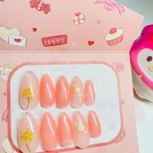 Söta rosa press-on naglar med blommor - Supergulliga press-on naglar i mandelform med skimrande rosa och ljusrosa bas. Några naglar har handmålade gula och vita blommor för extra söt detalj. Perfekt för dig som vill ha en snabb och fin nagel-look utan att gå till salong.