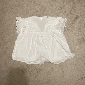 Vit blus med volang från Gina Tricot - Supersöt vit blus från Gina Tricot med v-ringning och volangdetaljer på ärmarna. luftig, broderade partier upptill och är tillverkad i ett lätt, mjukt tyg. 