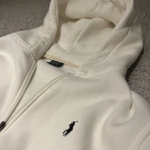 Vit hoodie från Polo Ralph Lauren - Snygg vit hoodie från Polo Ralph Lauren med dragkedja och klassisk broderad logga på bröstet. Perfekt för en chill och stilren look. Tillverkad i mjuk bomull för extra komfort. 