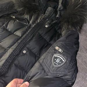 Svart dunjacka Rockandblue med fuskpäls - Snygg svart dunjacka från Rockandblue med quiltade detaljer och stor huva med fuskpäls. Jackan har flera dragkedjefickor, Rockandblue-logga på ärmen och en schysst, puffig look. Perfekt för kalla dagar när du vill hålla stilen.