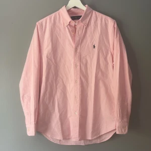 Rosa skjorta från Ralph Lauren S - Klassisk rosa skjorta från Ralph Lauren med button down-krage och broderad logga på bröstet. Skjortan är långärmad, har normal passform och är tillverkad i bomull. Perfekt för dig som gillar stilrena och tidlösa plagg. Hör av dig vid frågor och funderingar 