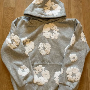 Grå hoodie från Denim Tears med tryck - Säljer en grå hoodie från Denim Tears med unika vita bomullsliknande tryck över hela tröjan. Tröjan har huva och magficka, samt ribbade muddar vid ärmslut och nederkant. Perfekt för dig som gillar streetwear och vill sticka ut med en cool design.