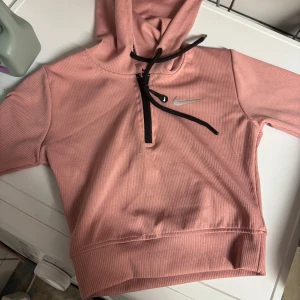 Rosa hoodie från Nike med half zip - Säljer en snygg rosa hoodie från Nike med svart snörning och half zip framtill. Tröjan har huva, långa ärmar och är tillverkad i ett ribbat material. Perfekt för dig som gillar sportig och avslappnad stil. Finns även byxa i separat annons. Storlek S men passar även xxs 