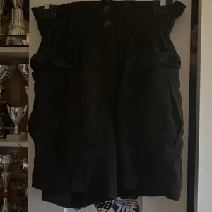 Svarta högmidjade shorts strl 36 - Svarta shorts med hög midja och bred resår upptill. Stora fickor på sidorna och knappar framtill ger en cool och avslappnad vibe. Perfekta för varma dagar när du vill ha både stil och komfort.