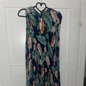 Blommig långklänning i marinblått - Snygg långklänning med marinblå bas och stora blommor i orange, vitt och grönt. Ärmlös modell med avslappnad passform, perfekt för varma dagar. Lätt och luftig, passar dig som gillar färgstarka mönster och somrig stil.