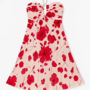 Plisserad blommig kortklänning helt ny - Supersöt kortklänning med plisserat tyg i ljusbeige och stora röda blommor. Axelbandslös modell med hjärtformad byst och lätt utsvängd kjol. Perfekt för dig som vill sticka ut med ett romantiskt och färgstarkt plagg. Helt ny och oanvänd 