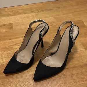 Svarta pumps med kedjedetalj - Snygga svarta pumps med spetsig tå och hög klack. Skorna har en cool silverfärgad kedja som rem runt hälen och är gjorda i mockaliknande material. Perfekta för dig som vill sticka ut med en edgy detalj till outfiten.