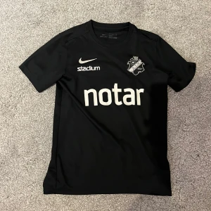 Svart AIK fotbollströja Nike - AIK fotbollströja i svart med vita tryck, inklusive klubbmärke, Nike-logga och sponsorer som Notar, Fabege och SVEA. Tillverkad i lätt och ventilerande Dri-FIT-material, perfekt för träning eller match. Klassisk passform med rund hals och korta ärmar.