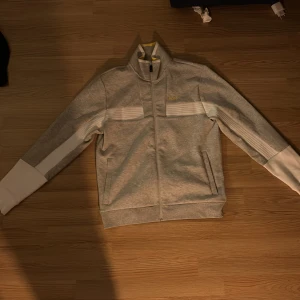 Grå tröja från BOSS med vita detaljer - Snygg grå tröja från BOSS med vita paneler på ärmarna och ribbade detaljer över bröstet. Jackan har hel dragkedja, ståkrage och diskret BOSS-logga i gult på bröstet. 