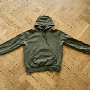Olivgrön hoodie från Polo Ralph Lauren - Snygg olivgrön hoodie från Polo Ralph Lauren med klassisk broderad logga på bröstet. Tröjan har huva med snörning, känguruficka och långa ärmar. Tillverkad i mjuk bomull för en skön och avslappnad look.