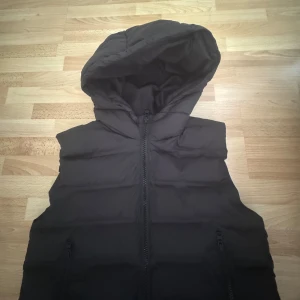 Svart dunväst med huva från Zara - Säljer en svart dunväst från Zara i storlek 164. Västen har en stor, mysig huva och två fickor med dragkedja framtill. Quiltad design med breda kanaler och hel dragkedja. Perfekt att slänga över en hoodie för extra värme. Använd en gång. Storlek 164, men passar xs/s också. Längd 70cm, bröst 104cm.