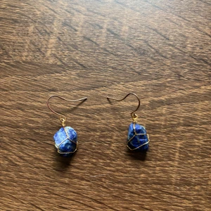 Blå sten örhängen med guldtråd - Unika örhängen med blå, oregelbundna stenar insvepta i tunn guldtråd. Hängande modell med krok, perfekt för dig som gillar boho eller naturinspirerad stil. Stenarna har olika blå nyanser och detaljer i guld som ger en cool kontrast.