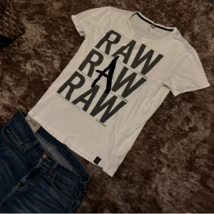 Vit G-Star RAW t-shirt med tryck - Pris kan diskuteras vid snabb affär. Vit t-shirt från G-Star RAW med stort grafiskt RAW-tryck i svart och grått på framsidan och texten 'THE ART OF RAW' på ryggen. Klassisk rund hals och korta ärmar. Tillverkad i mjuk bomull för en skön känsla och avslappnad känsla.