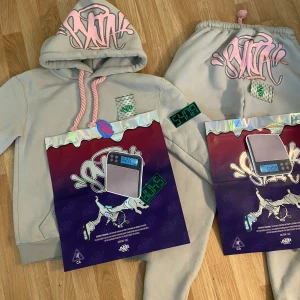 Syna World Set (Hoodie + Sweatpants) - Säljer ett komplett Syna World set i färgen ljusgrå med rosa detaljer – extremt eftertraktat och helt oanvänt!  - Setet består av hoodie + byxor -Fet design med rosa graffiti-logga, tryckt patch och detaljerad artwork - Aldrig använt – i helt nytt skick -Kommer med original förpackning och tags