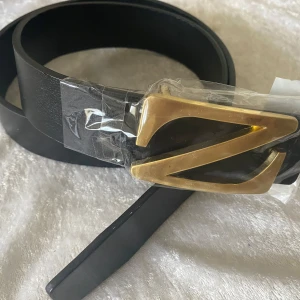 Svart bälte med guldspänne Z - Stilrent svart bälte i skinn med ett stort, guldigt Z-format spänne som verkligen sticker ut. Bältet har en klassisk, rak form och passar perfekt för dig som vill ha en snygg detalj till outfiten. Enkel design utan mönster, vilket gör det lätt att matcha. Helt ny , Kolla bild 2 , sälj rabatt pris 