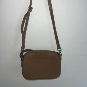 Brun axelväska från Trussardi - Snygg brun axelväska från Trussardi i strukturerat skinn. Väskan har justerbar axelrem, silverfärgade metalldetaljer och dragkedja upptill. Perfekt storlek för det viktigaste och stilren design med diskret logga framtill.