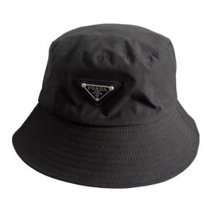 Svart bucket hat från Prada - Fullända din look med denna ikoniska svarta Prada bucket hat. Tillverkad av högkvalitativa material och prydd med den klassiska triangulära Prada-logotypen i silver, är denna hatt både stilren och praktisk. Perfekt för att skydda dig mot solen eller bara för att lägga till en extra touch av lyx till din outfit 