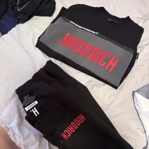 Svart sweatshirt från Hoodrich - Svart sweatshirt från Hoodrich med grå panel över bröstet och stor röd logga framtill. Crewneck-modell med en clean och sportig vibe, perfekt för dig som gillar streetwear. Materialet känns mjukt och bekvämt, troligen bomull eller polyester.