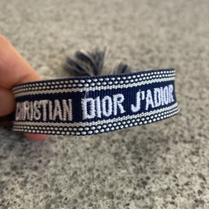 Blått armband Christian Dior J'Adior - Snyggt blått tygarmband från Christian Dior med texten 'J'Adior' broderad i vitt. Armbandet har justerbara tofsar i ändarna och ett coolt, vävt mönster längs kanterna. Perfekt accessoar för att lyfta din outfit med en lyxig touch.