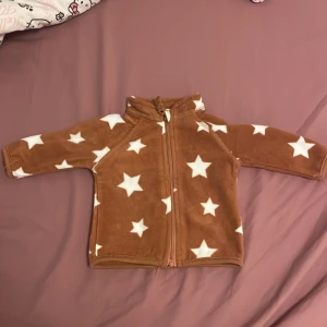 Brun fleecejacka med stjärnor från Ergee - Supermjuk brun fleecejacka från Ergee i storlek 56, täckt med vita stjärnor. Jackan har dragkedja framtill och långärmade ärmar. Perfekt för att hålla de minsta varma och mysiga under kyliga dagar.