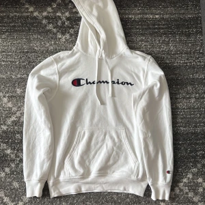 Vit Champion hoodie med broderad logo - Säljer en clean vit hoodie från Champion i storlek S. Tröjan har broderad Champion-logo på bröstet, känguruficka och dragsko i huvan. Perfekt för dig som gillar streetwear och enkel stil. Skön bomullsmix och långa ärmar.