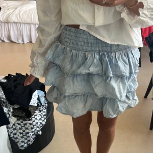 Volangkjol i ljus denim från Ginatricot  - Supersöt kort volangkjol i ljusblå denim från Ginatricot . Kjolen har bred smock i midjan, flera lager volanger och dragkedja bak. .