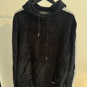 Svart hoodie från Manière De Voir - Svart hoodie i mjuk sammet från Manière De Voir med gråa och vita ränder längs ärmarna. Tröjan har huva med snörning och en stor magficka framtill. Perfekt för dig som gillar en chill och stilren look.