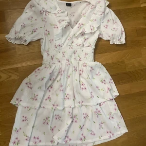 Vit blommig kortklänning med volang - Supersöt vit kortklänning med rosa blommor och gröna blad. Klänningen har volangdetaljer vid halsringningen, v-ringad urringning och puffiga korta ärmar med resår. Smockad midja och dubbla lager i kjolen ger en romantisk vibe. Aldrig använd