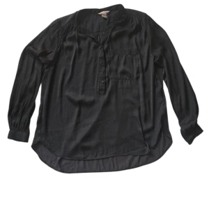 Svart prickig henley från H&M - Svart henley-skjorta från H&M i storlek 44. Skjortan har diskreta små prickar, lång ärm, bröstficka och knappar framtill. Materialet är glansigt och känns mjukt mot huden, perfekt för en chill och stilren look.