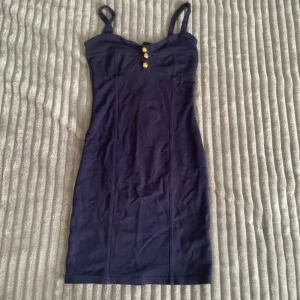 Mörkblå tight klänning med guldknappar från Art Hole - Säljer en mörkblå tight klänning från bikbok med smala axelband och tre dekorativa guldknappar framtill. Klänningen har markerad byst och dragkedja i ryggen. Perfekt för dig som gillar en stilren och figurnära look.