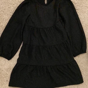 Svart volangklänning från Lindex Kids - Säljer en svart klänning från Lindex Kids i storlek 134. Klänningen har lång ärm, flera volanglager och en lätt strukturerad yta i tyget. Perfekt för dig som gillar en enkel men stilren look.