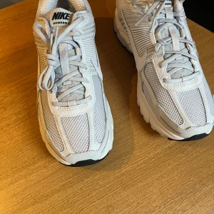 Nike Vomero 5 sneakers i ljusgrått - Säljer ett par Nike Vomero 5 sneakers i ljusgrått med mesh och syntetpaneler. Skorna har snörning, rund tå och en chunky sula för extra komfort. Perfekta för dig som gillar stilrena och sportiga sneakers.